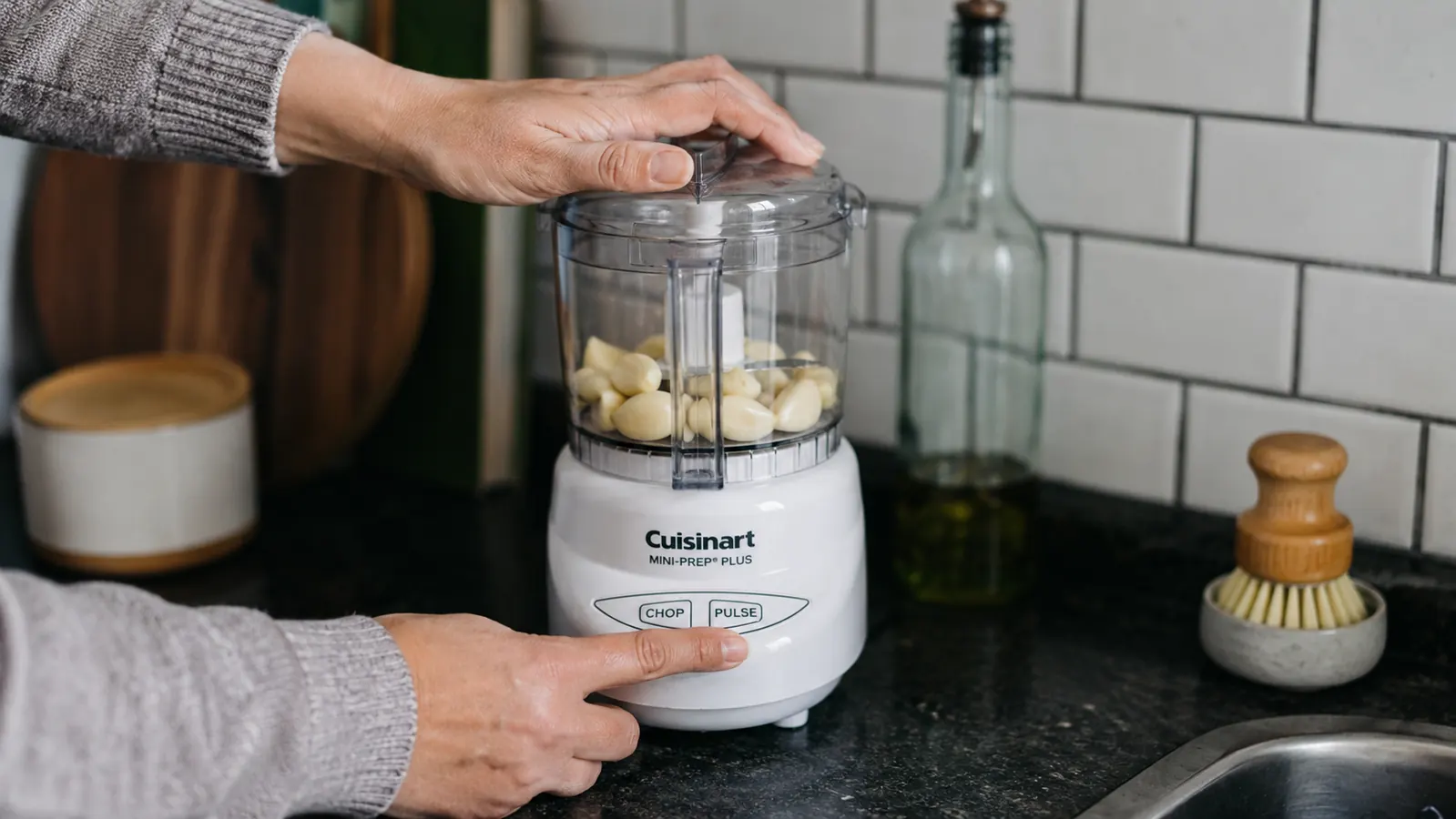 Hand pressing the pulse button on a Cuisinart Mini-Prep Plus while garlic cloves sit in the clear bowl